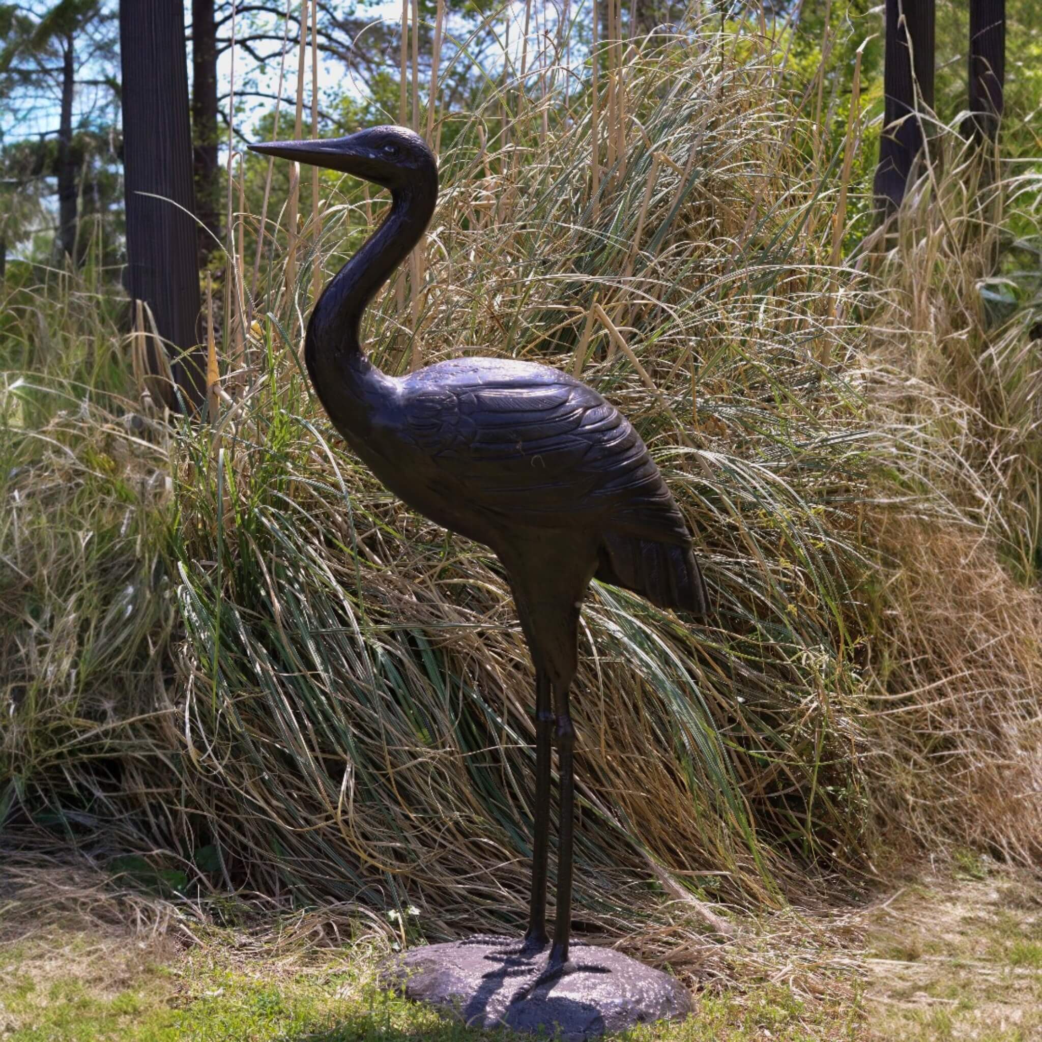 Life-size cast aluminum heron sculpture 62 inches tall standing in ornamental grass with trees in background