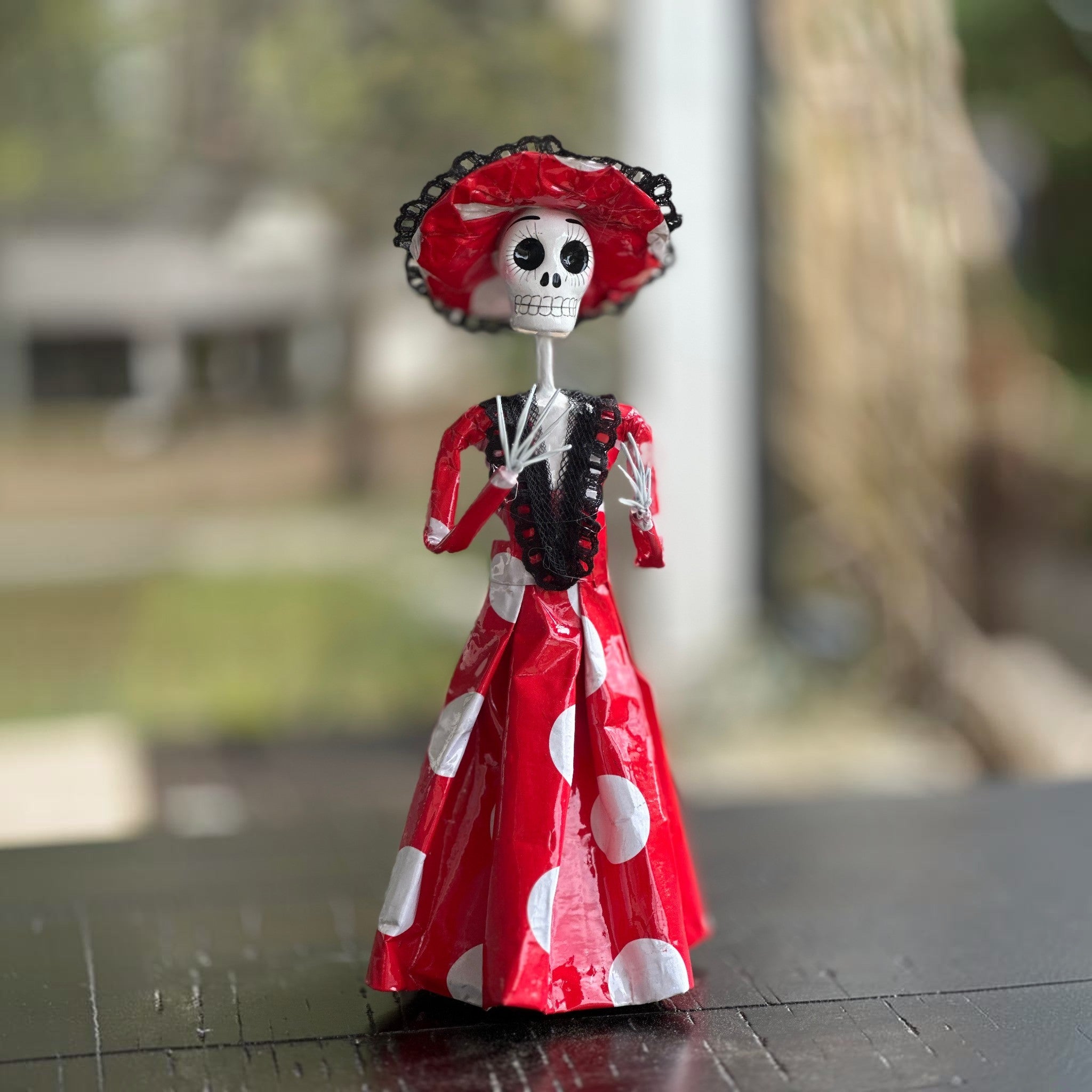 Hand-painted Mexican Catrina figurine in red polka dot dress and lace hat on a wooden porch, traditional Día de los Muertos folk art