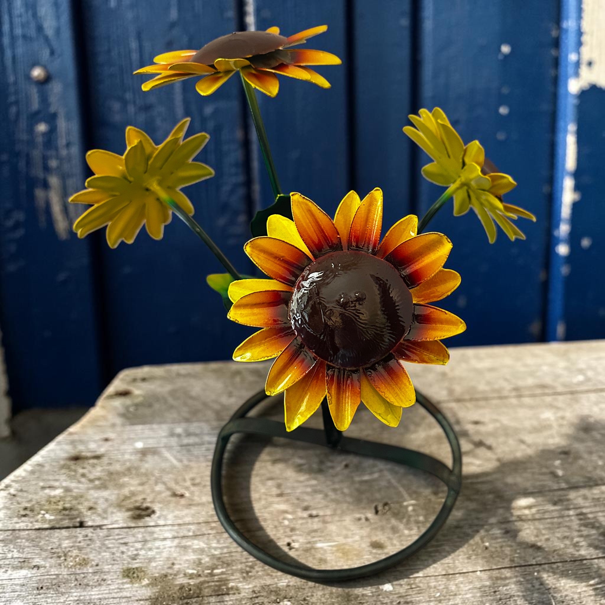 Hand-painted metal sunflower garden sculpture with yellow and orange petals on weathered grey wood, blurred deep navy painted door background, rustic outdoor folk art