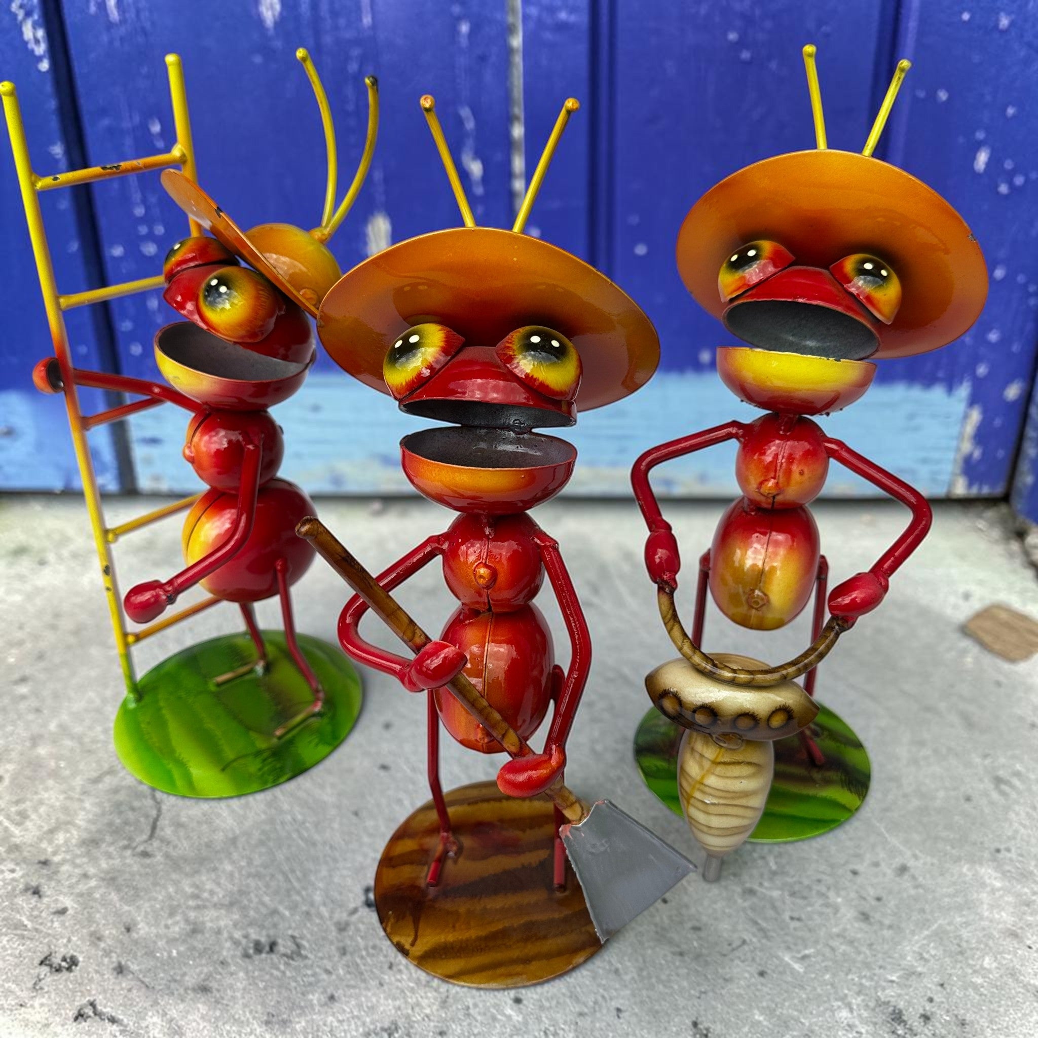 Three Mexican folk art ant sculptures in red and orange with sombreros, carrying ladder, shovel, and auger drill against a bright royal blue weathered wood door