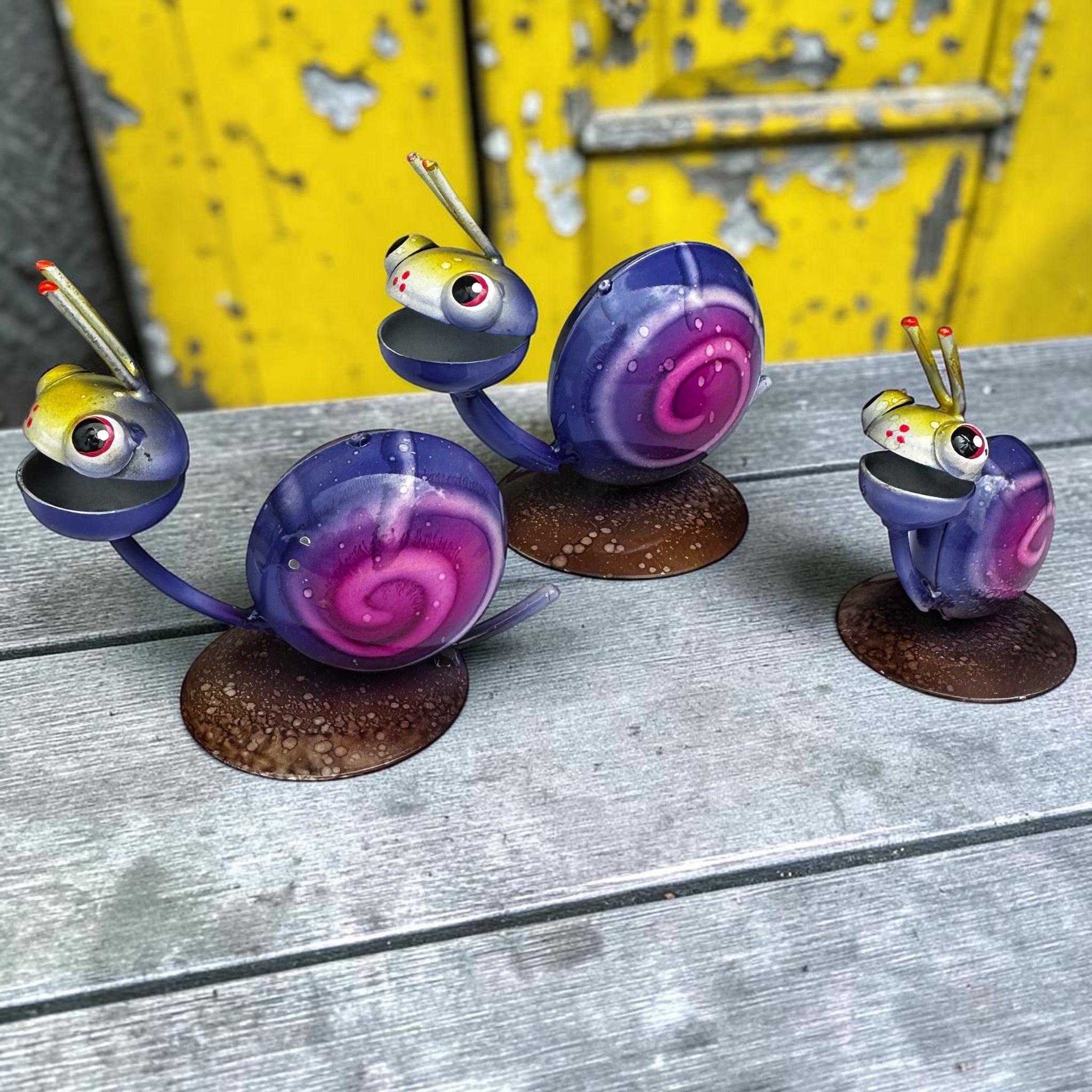 Three hand-painted Mexican folk art metal snail sculptures facing forward with expressive eyes and antennae, purple and pink shells against blurred yellow door background