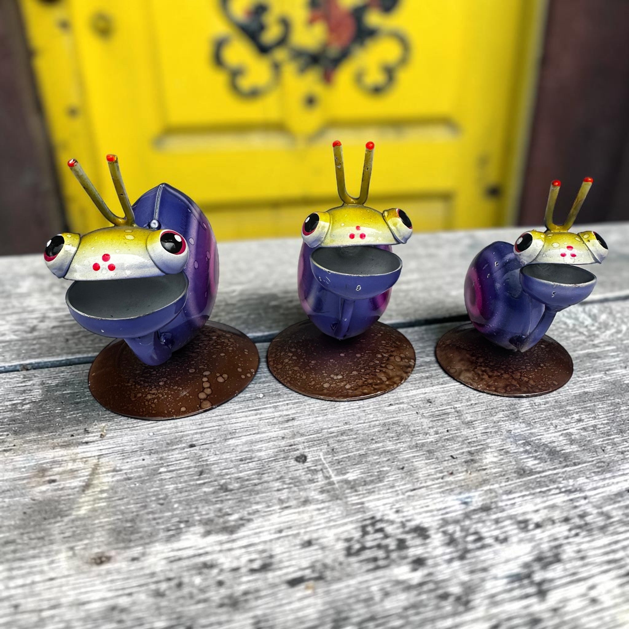 Side view of three Mexican folk art snail sculptures with purple swirl shells and yellow heads, marching across weathered grey wood with yellow door behind