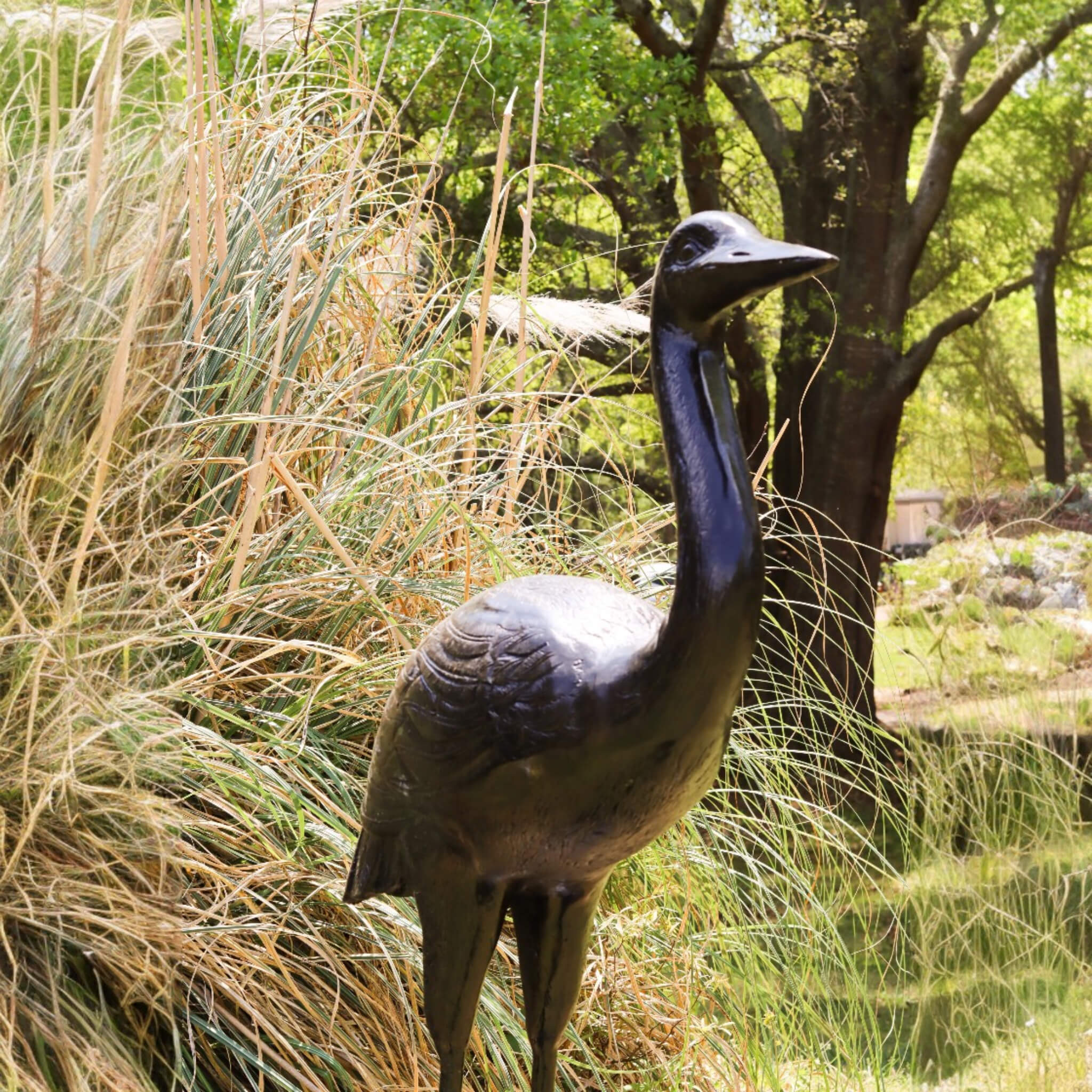 Cast aluminum heron garden sculpture close up showing dark patina feather detail with ornamental grass and pond in background