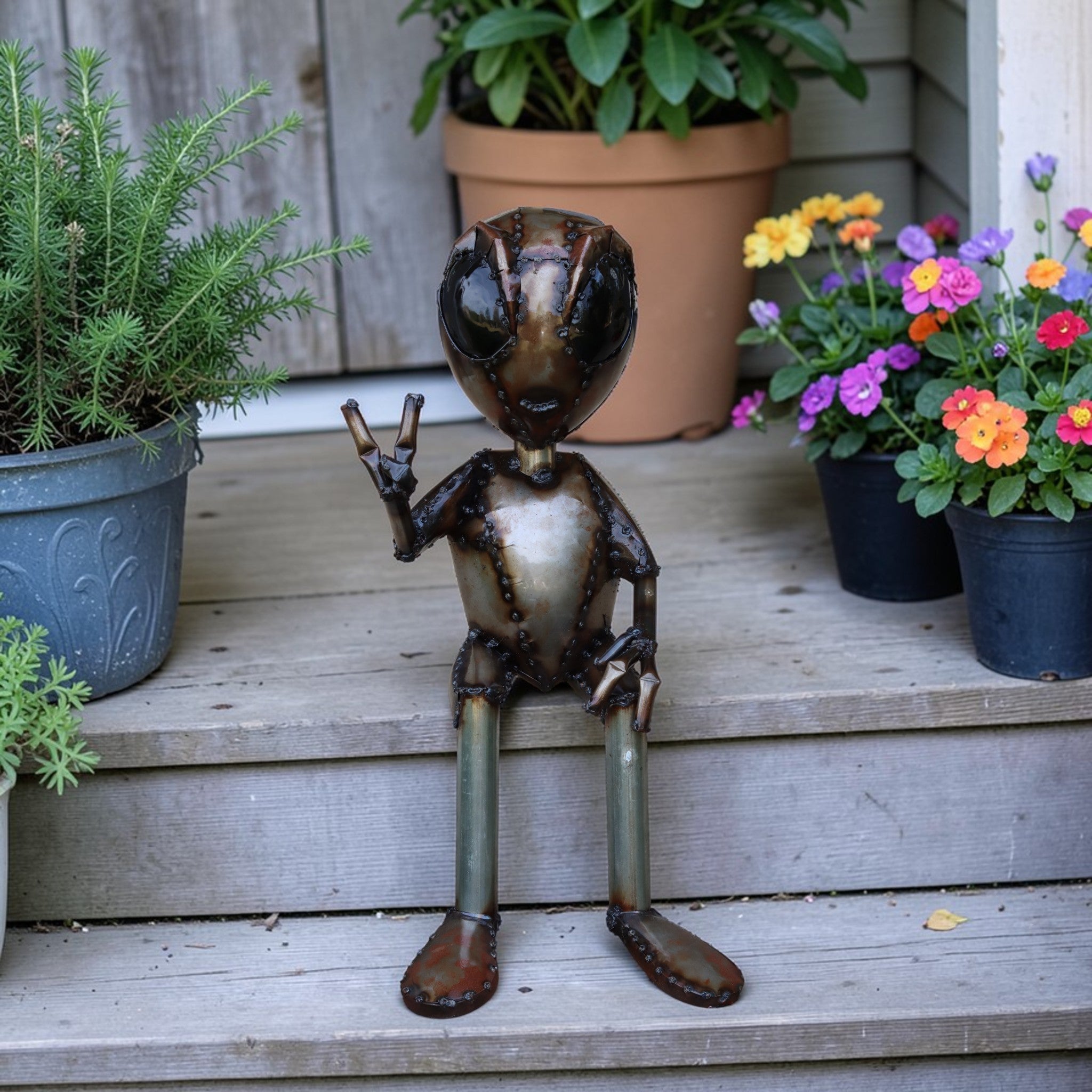 Metal alien garden sculpture making peace sign sitting on porch steps with potted plants