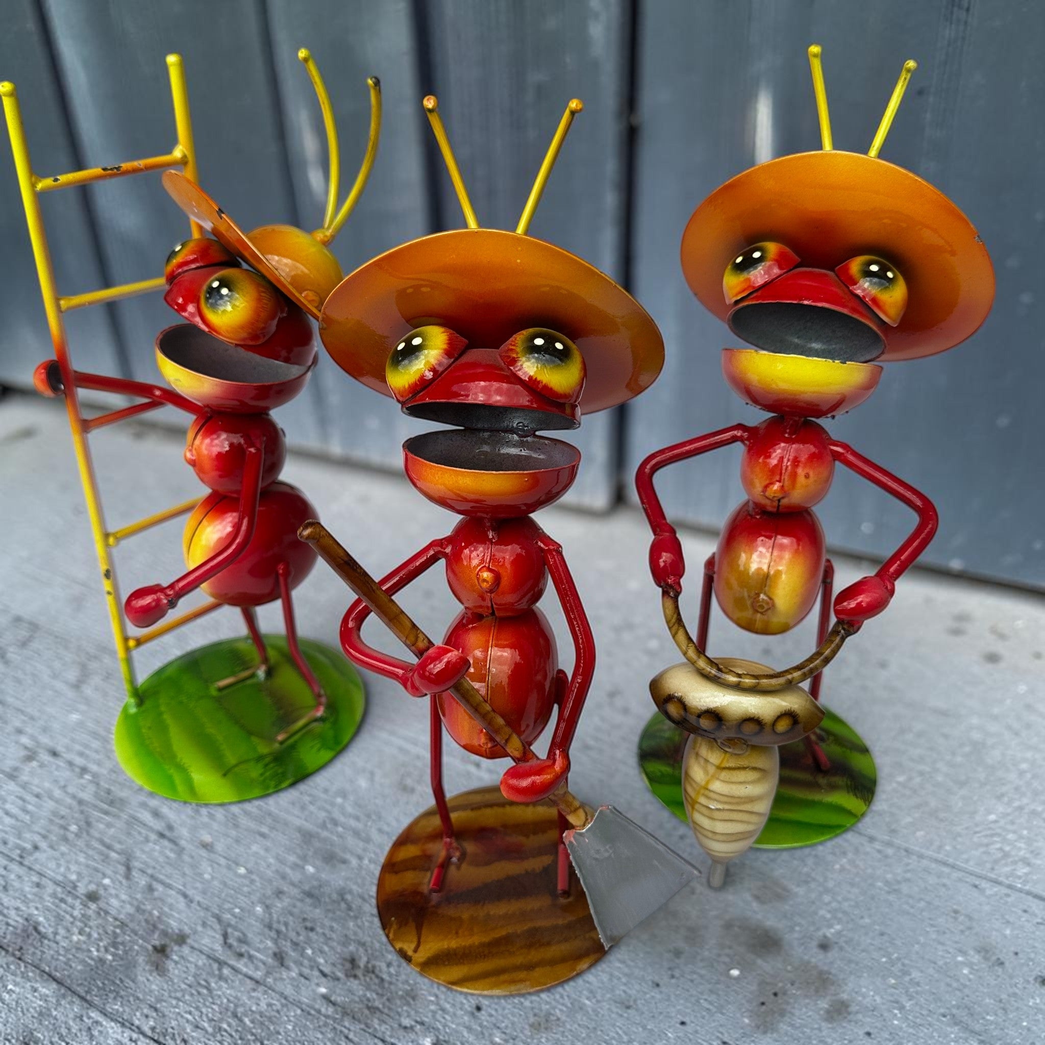 Three hand-painted Mexican folk art metal ant sculptures with red bodies and orange sombrero hats holding a ladder, shovel, and drill against a gray weathered wood background