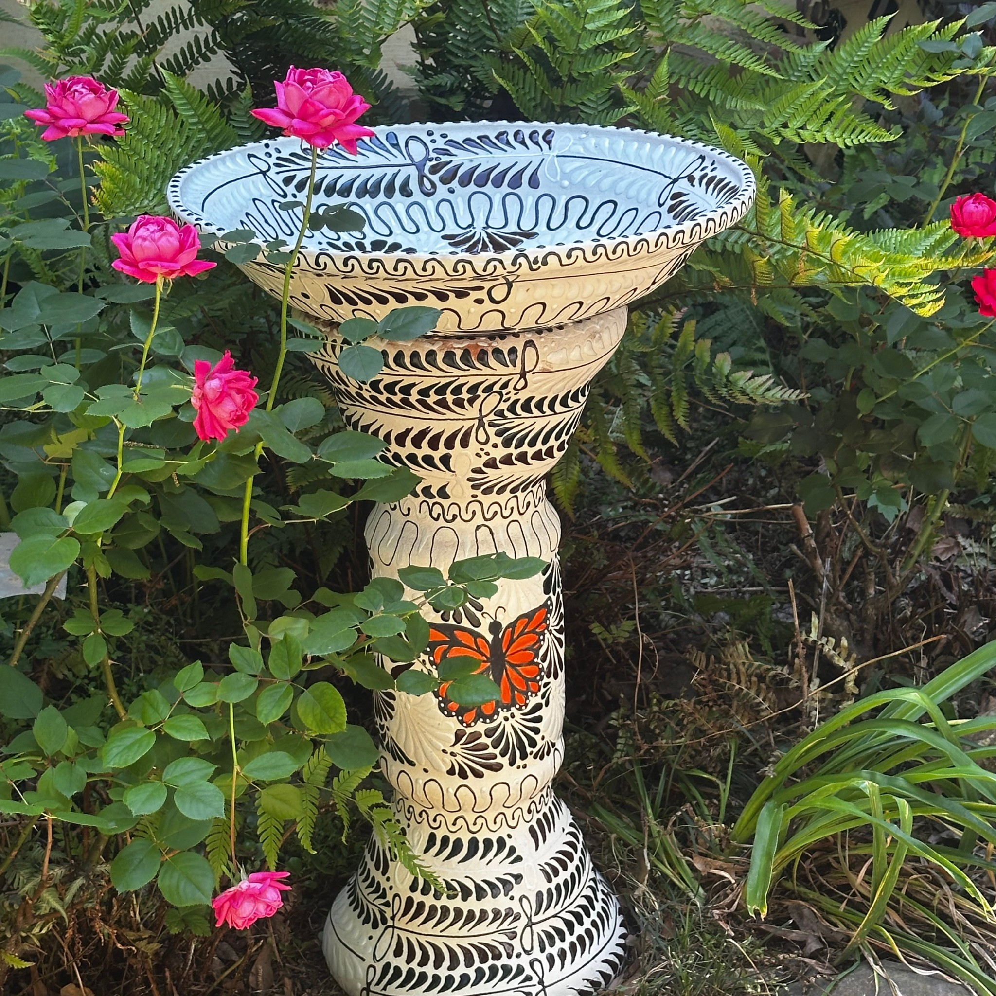 Hand-painted Mexican ceramic birdbath in white with black floral pattern and orange monarch butterfly, Tonalá folk art set in garden with pink roses