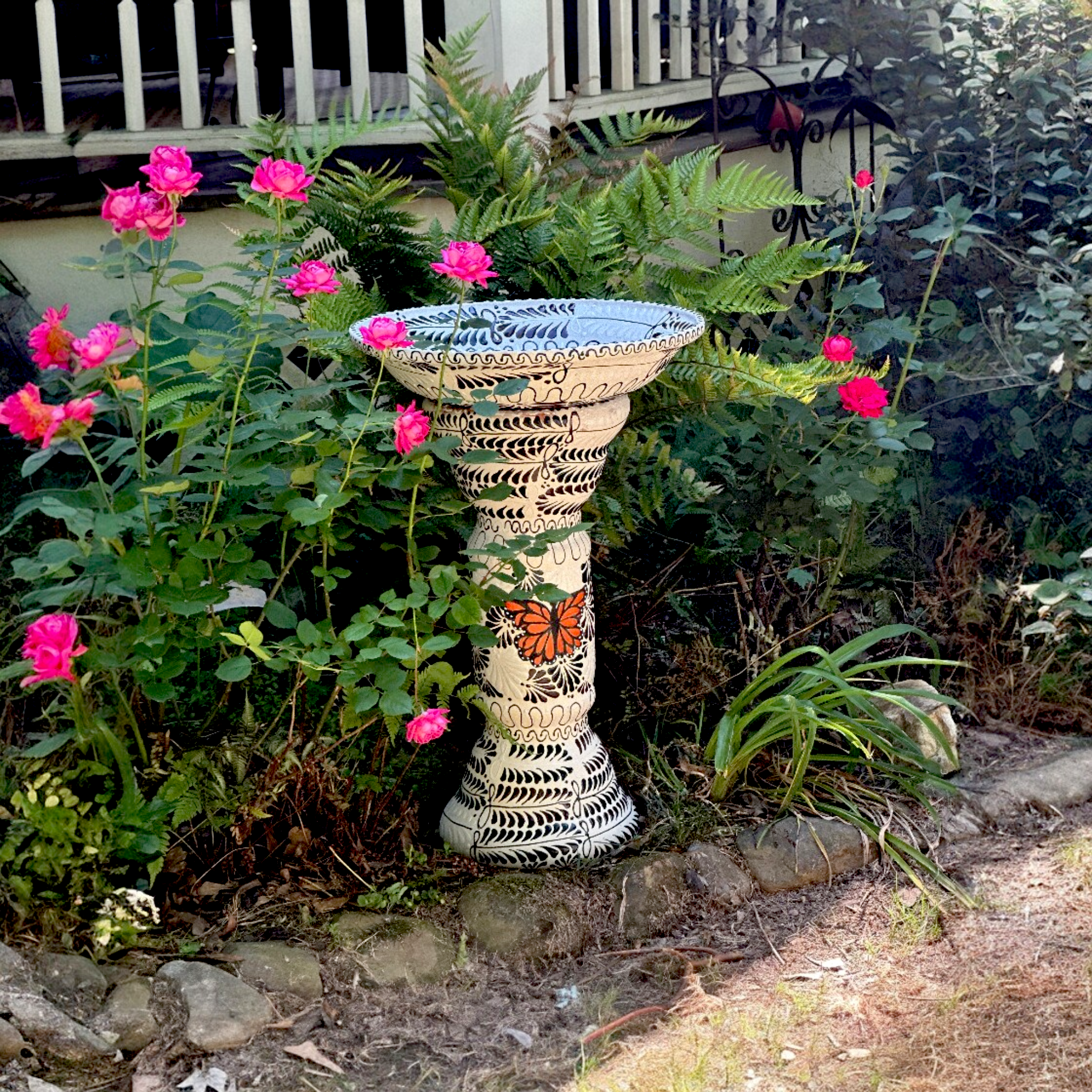 Hand-painted Mexican ceramic birdbath in white with black floral engobe pattern, orange monarch butterfly accent, hand-painted Tonalá folk art set in garden with pink roses and ferns