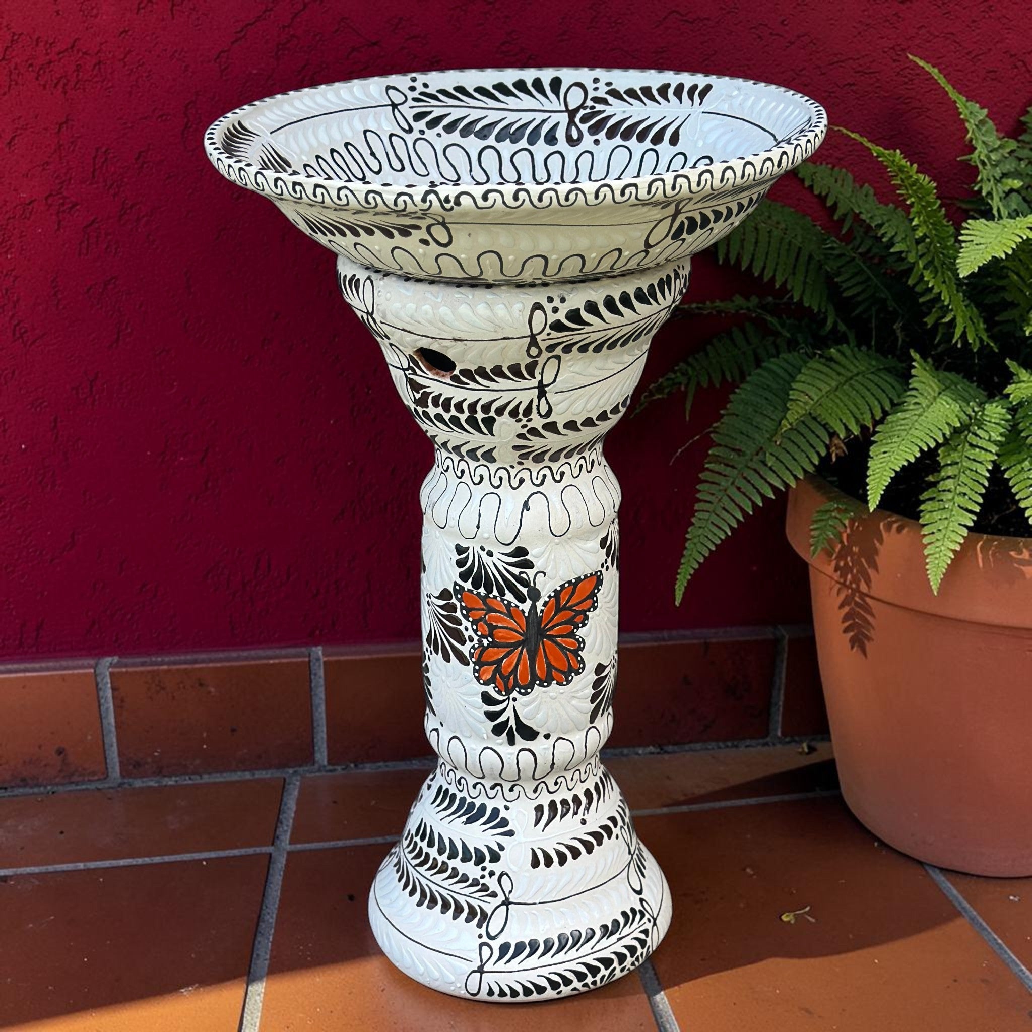 Hand-painted Mexican ceramic birdbath in white with black floral engobe pattern and orange monarch butterfly accent, Tonalá folk art, against red stucco wall with fern